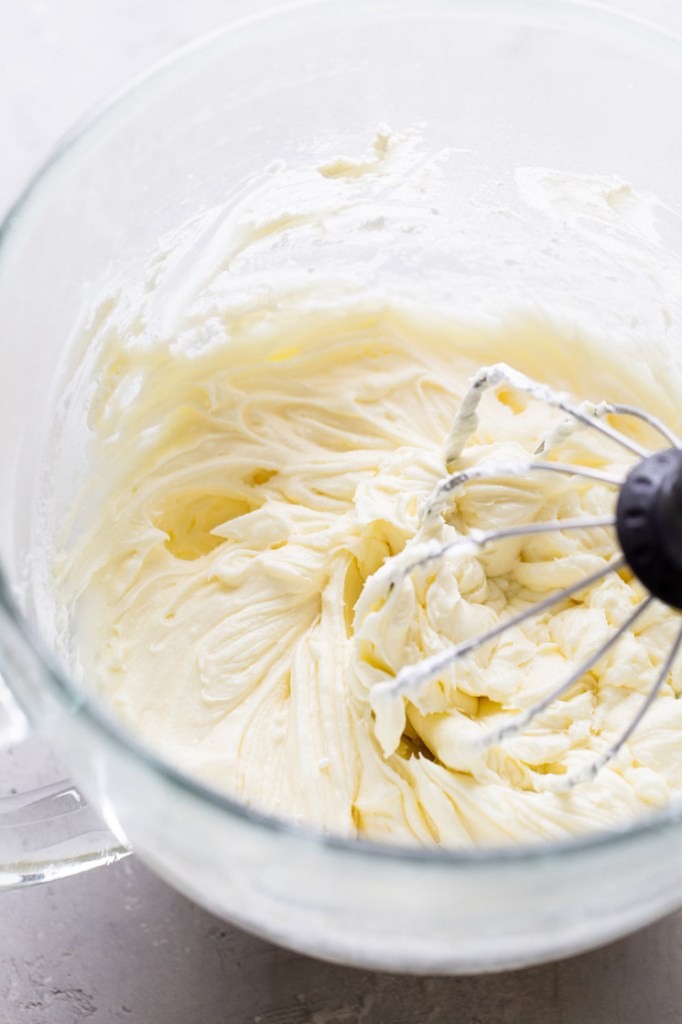 A glass mixing bowl filled with lemon cake frosting. A whisk attachment rests in the bowl. 