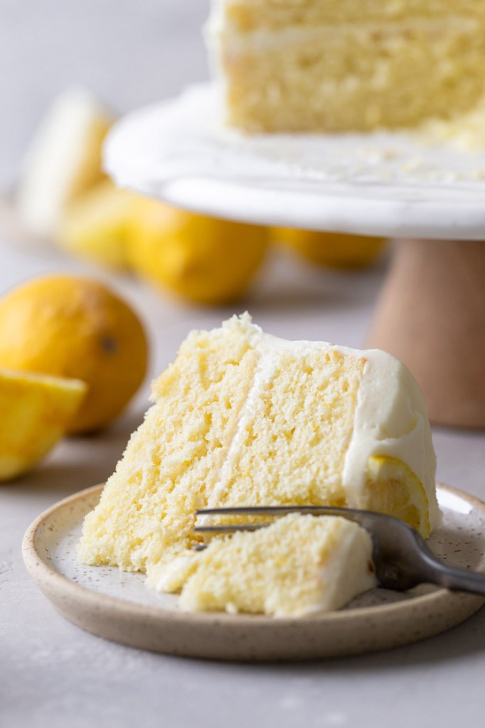 A slice of homemade lemon cake lying on its side on a speckled plate. A fork is digging into the slice. The rest of the cake is on a cake stand in the background. 