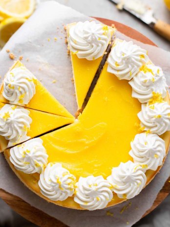 An overhead view of a lemon cheesecake topped with lemon curd and whipped cream. The cheesecake has been partially sliced and one slice has been removed.