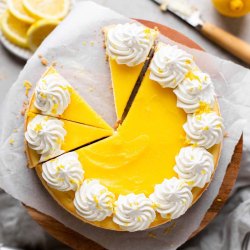 An overhead view of a lemon cheesecake topped with lemon curd and whipped cream. The cheesecake has been partially sliced and one slice has been removed.