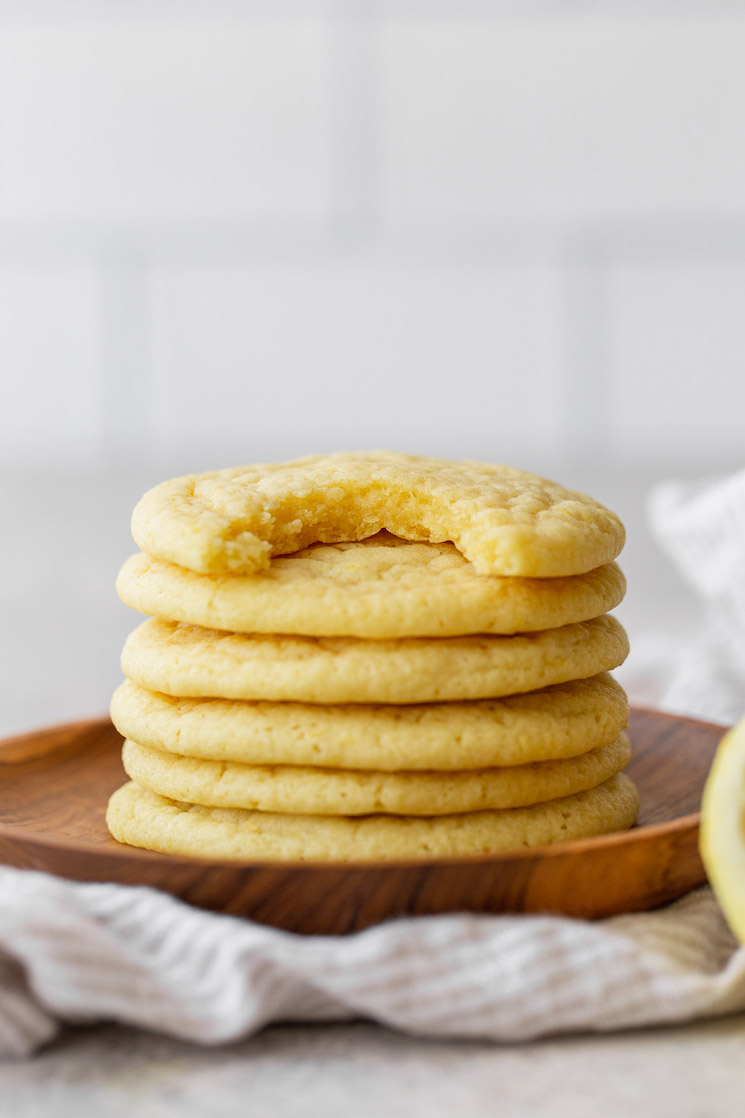 A stack of lemon cookies on a wooden plate, the top one has a bite taken out of it.