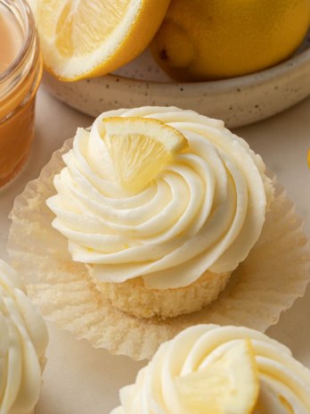 Lemon cupcakes topped with lemon buttercream and a lemon wedge. A bowl of lemons and jar of lemon curd are sitting next to the cupcakes.