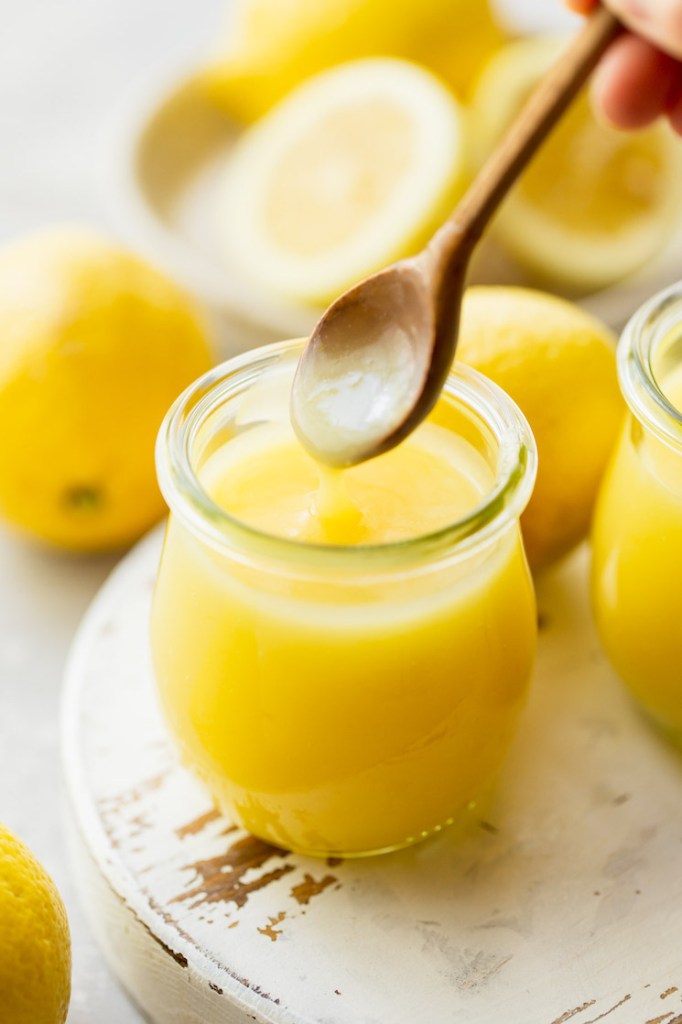 A wooden spoon scooping lemon curd out of a small glass jar.