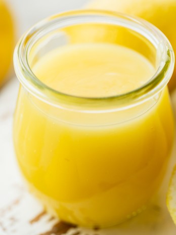 A small glass jar filled with lemon curd on top of a worn white wood surface.