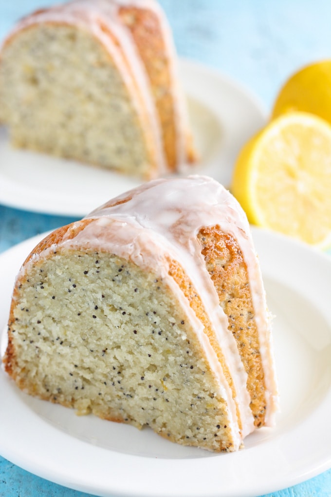 A slice of lemon poppy seed cake with glaze on a white plate. Lemon halves and another slice of cake rest in the background. 