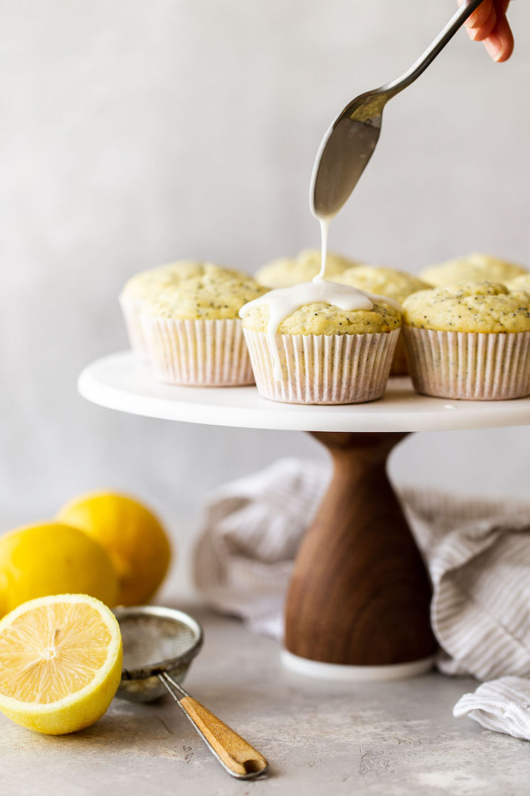 A marble cake stand with lemon poppy seed muffins being topped with an easy lemon glaze.