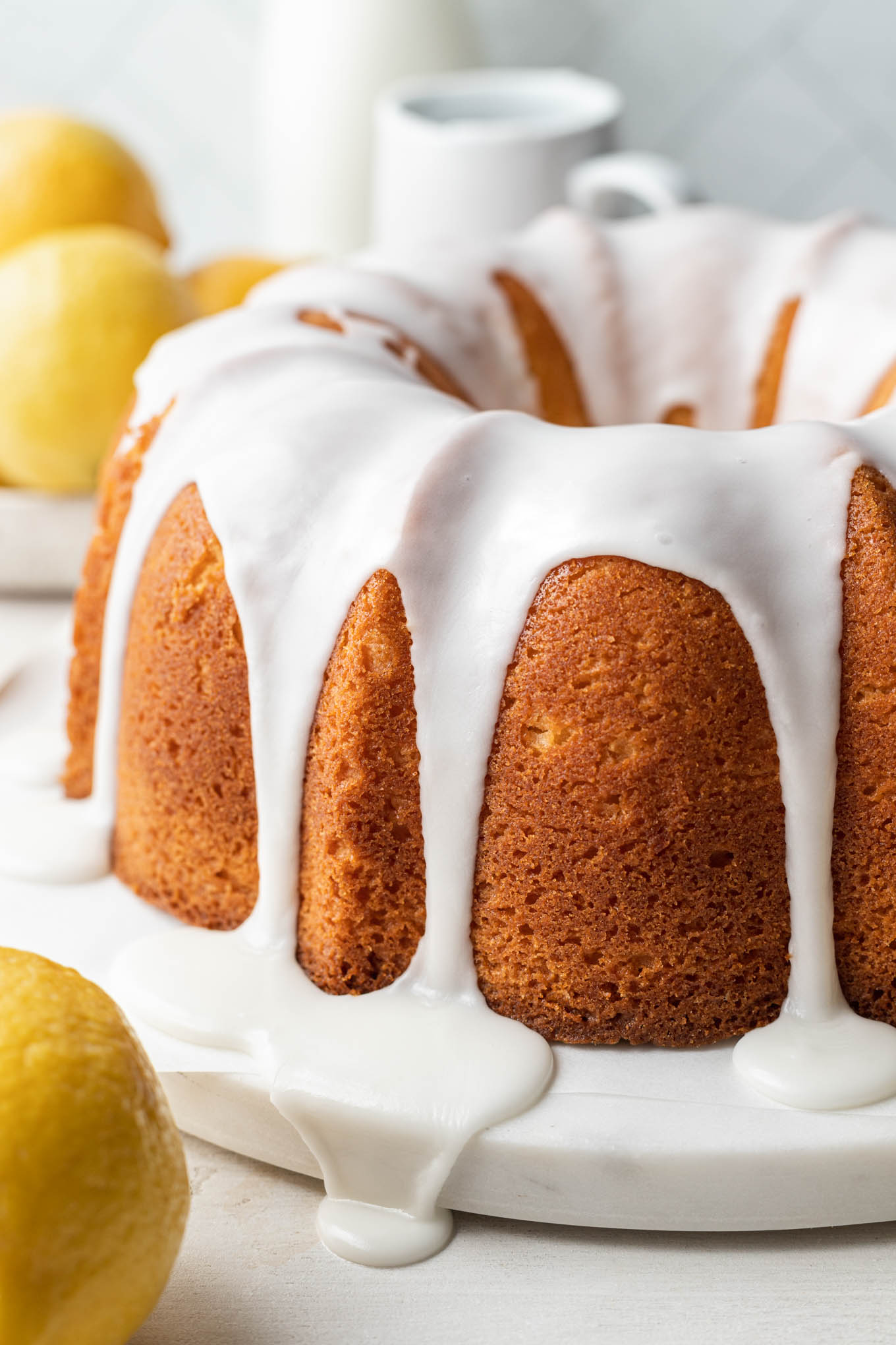 A glazed lemon pound cake on a white cake plate, with lemons in the foreground and background. 