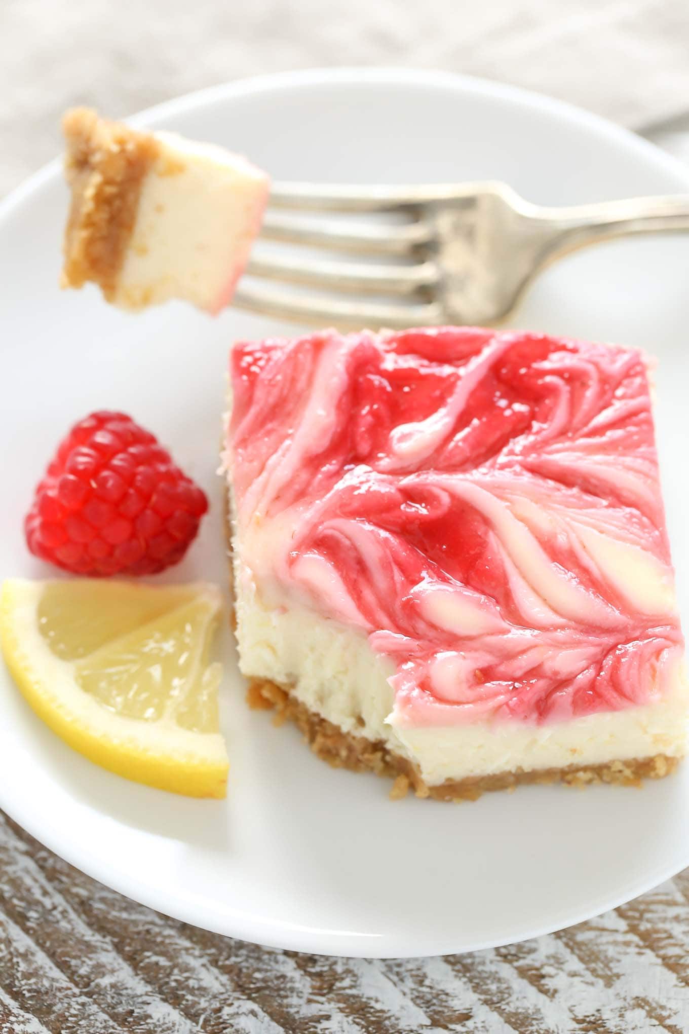 A raspberry swirl cheesecake bar on a white plate. A bite has been speared on a fork, which rests on the plate. There's also a lemon wedge and one fresh raspberry on the plate. 