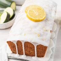 A loaf of lemon zucchini bread topped with two lemon slices. Some sliced zucchini rests in a small bowl on the side.