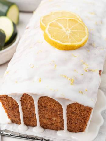 A loaf of lemon zucchini bread topped with two lemon slices. Some sliced zucchini rests in a small bowl on the side.