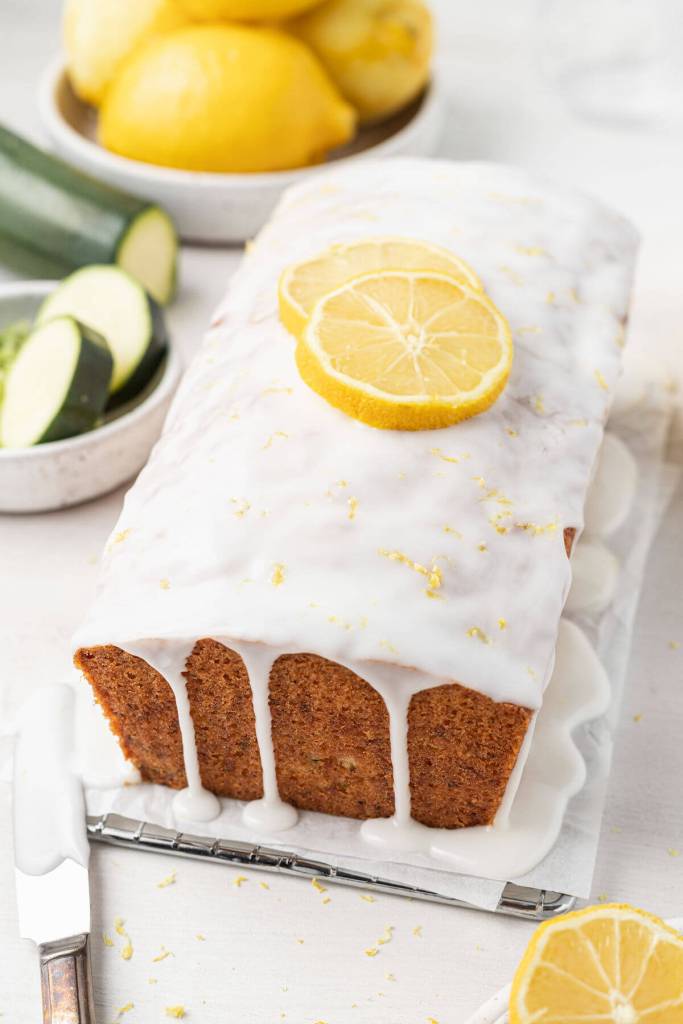 A loaf of glazed lemon zucchini bread on a parchment-lined wire rack. Dishes of sliced zucchini and lemons are in the background. 