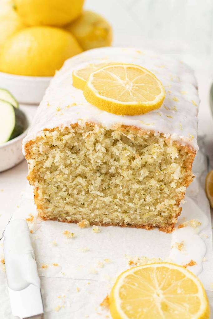 A loaf of glazed lemon zucchini bread, with a slice missing. A bowl of lemons rests in the background. 