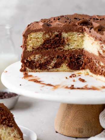 A side view of a homemade marble cake on a cake stand. A slice has been removed and has been plated. Milk jugs rest in the background.