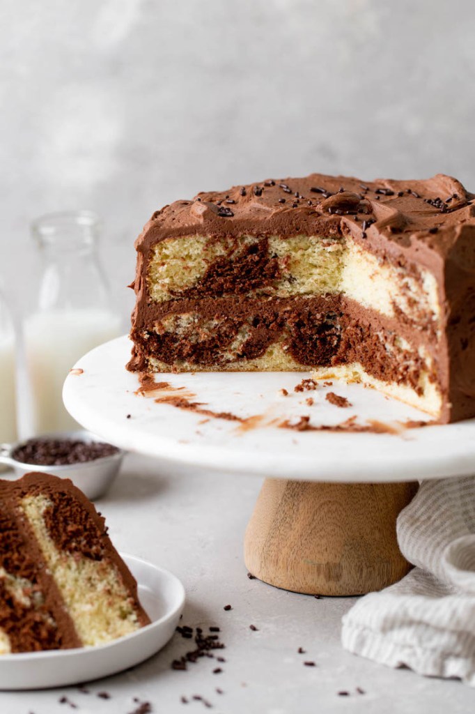 A side view of a homemade marble cake on a cake stand. A slice has been removed and has been plated. Milk jugs rest in the background. 