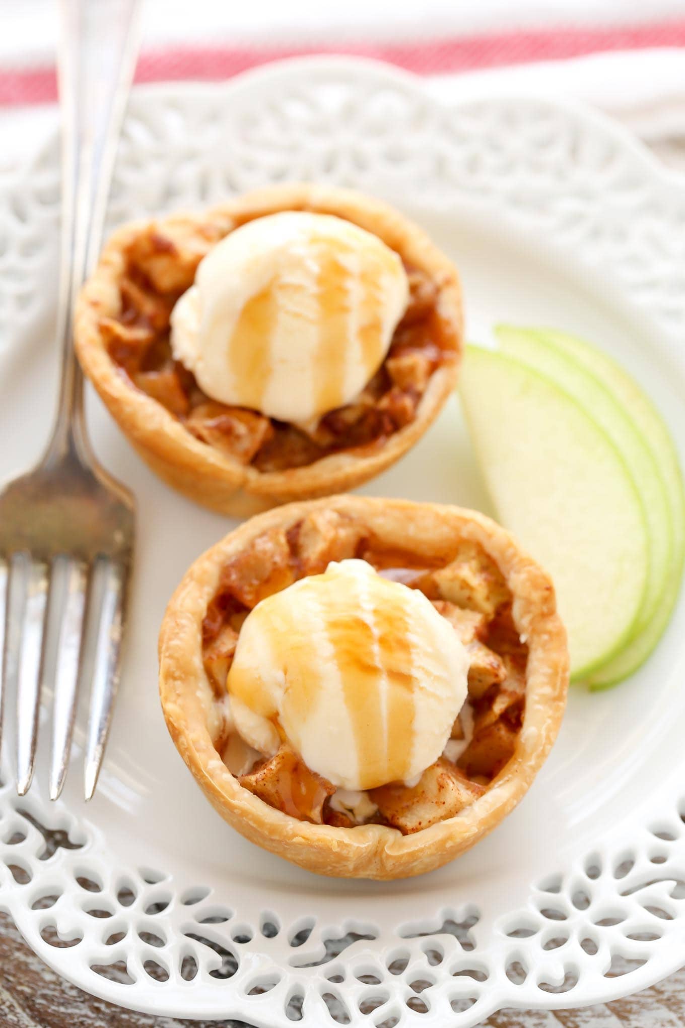 Two mini apple pies topped with a small scoop of ice cream and a drizzle of salted caramel sauce. A fork and a few slices of apples lay on the plate next to the pies.