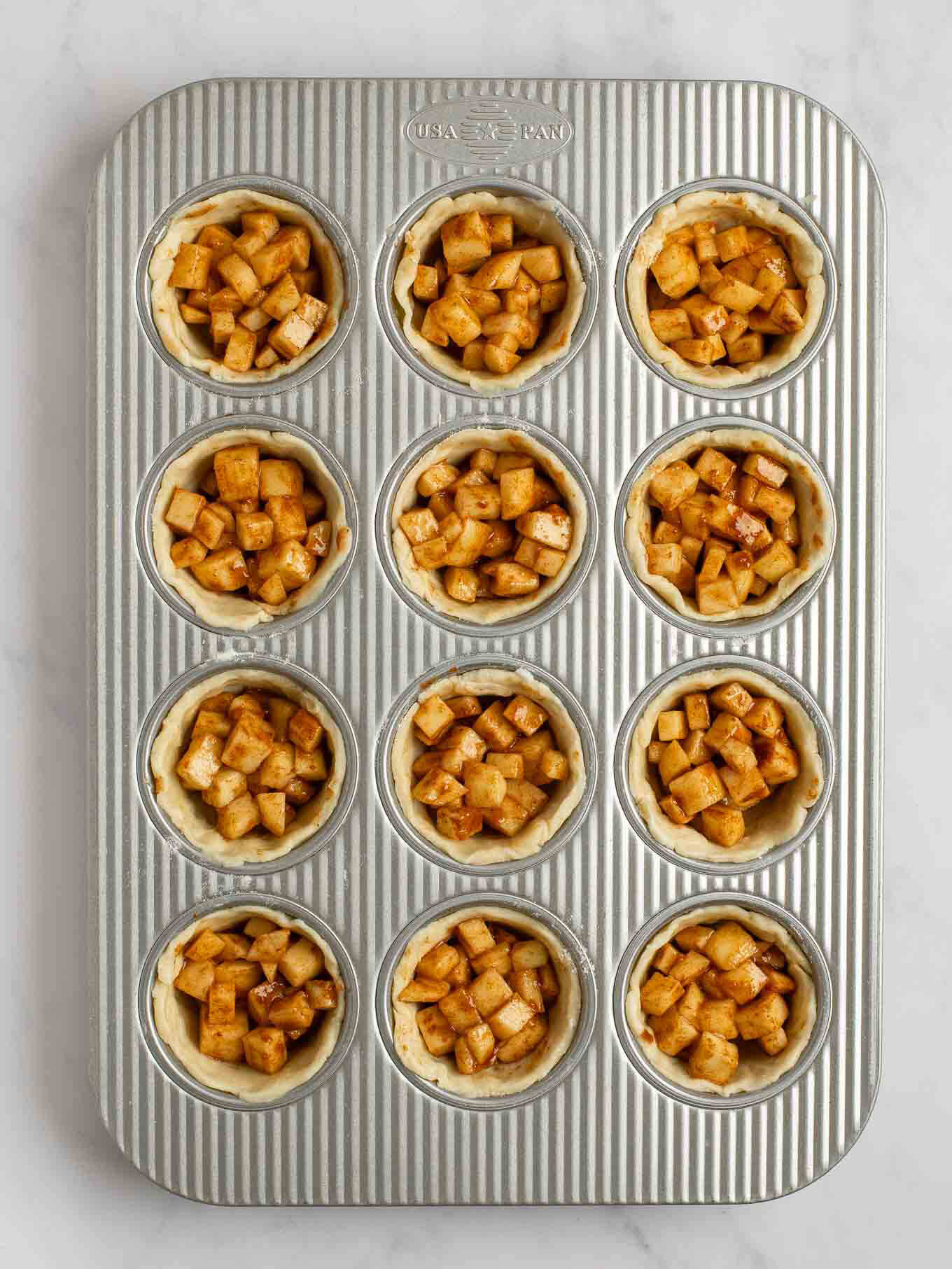 An overhead view of the apple pie filling evenly distributed between all of the pie crusts in a muffin pan.