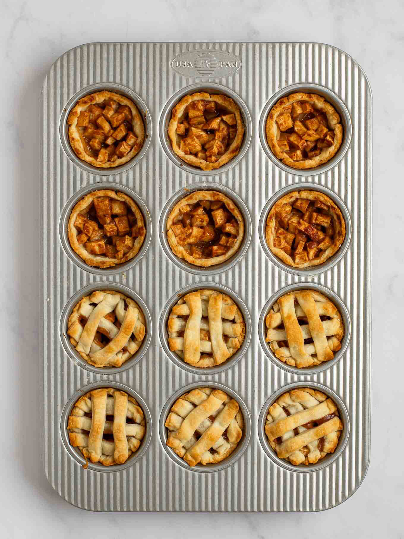An overhead view of the baked mini apple pies in a muffin pan. Half of the mini pies have a lattice pie crust topping.
