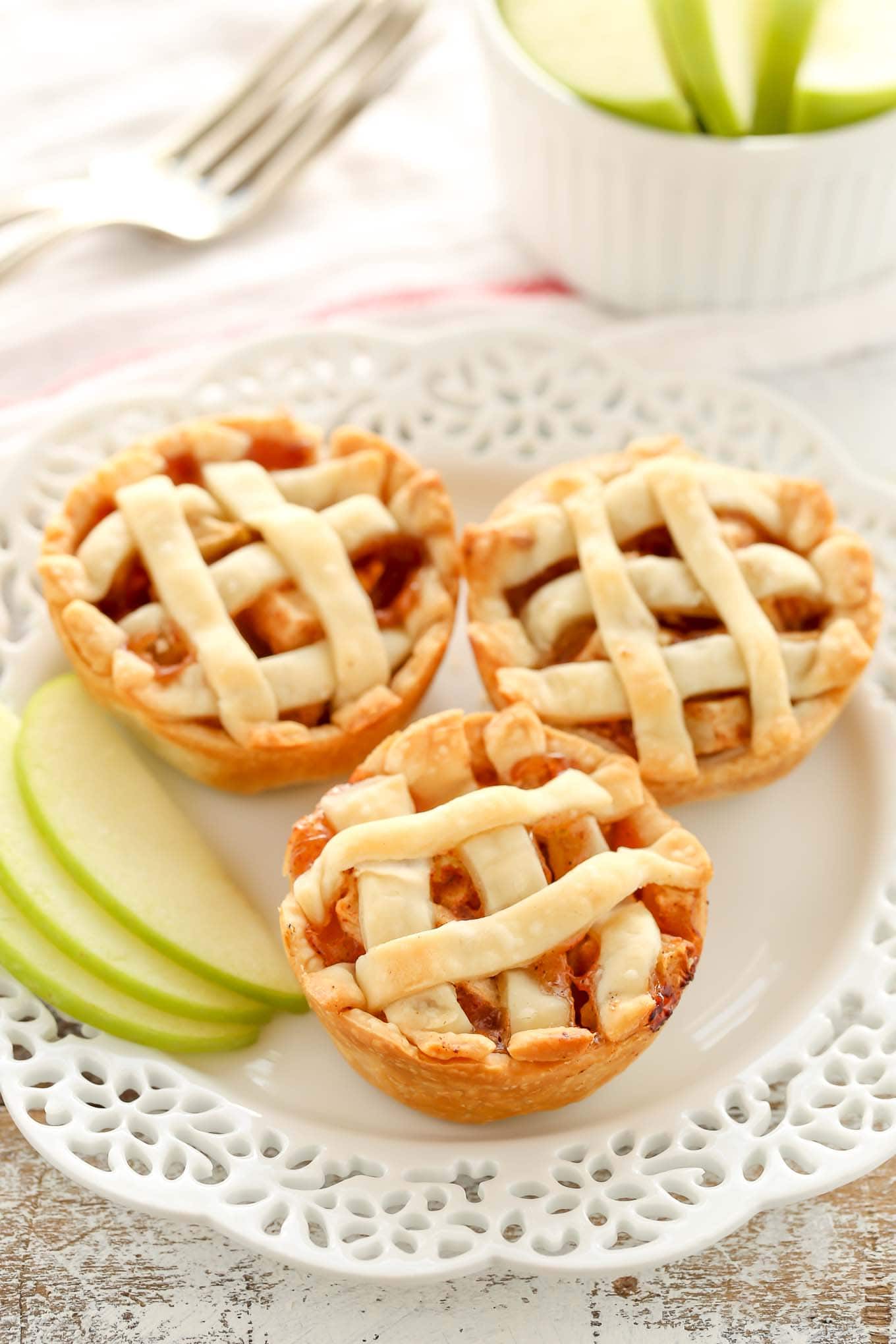 Three mini apple pies on a decorative white plate. A small container of sliced apples and a fork are in the background.