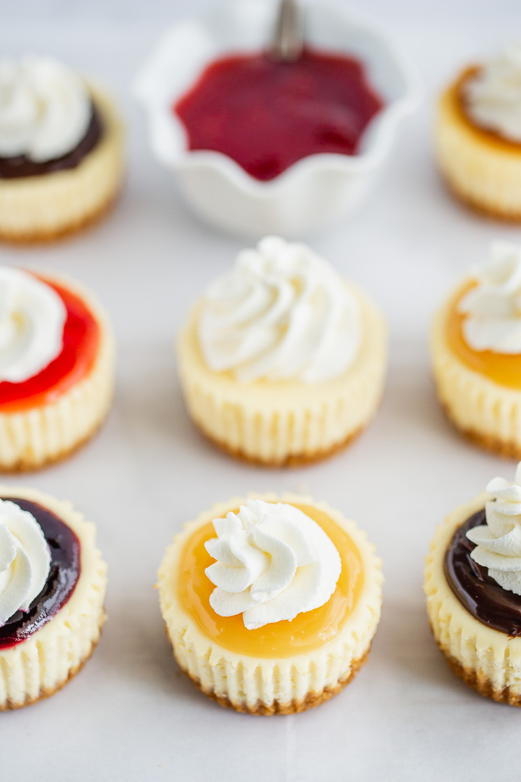 A piece of parchment paper lined with mini cheesecakes topped with different toppings and whipped cream.
