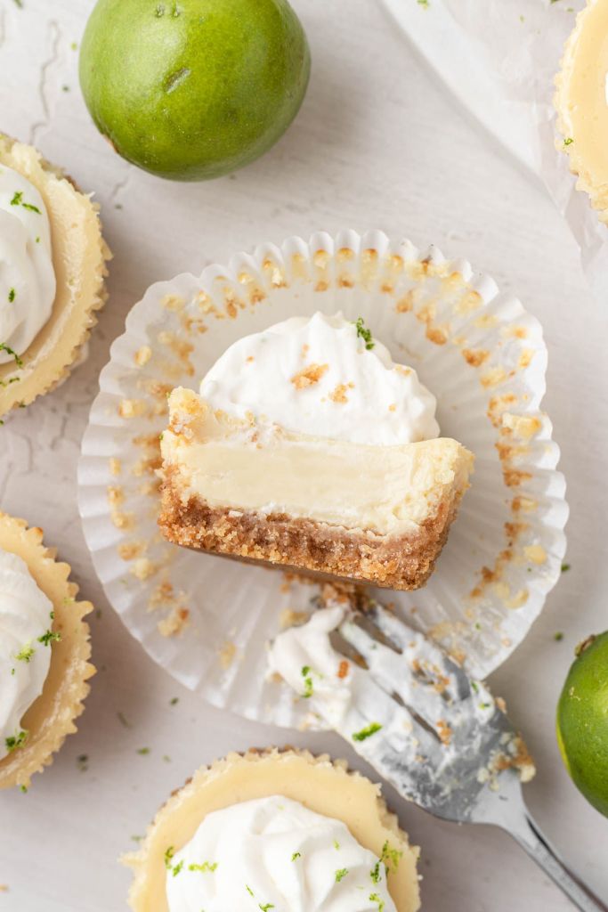 An overhead view of a mini key lime pie sitting on the wrapper. A bite has been taken out of it and more mini pies and key limes are around it.