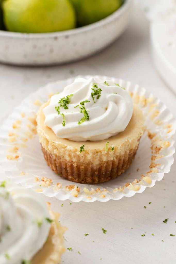 A mini key lime pie, with the muffin liner pulled down. A bowl of key limes is sitting in the background.