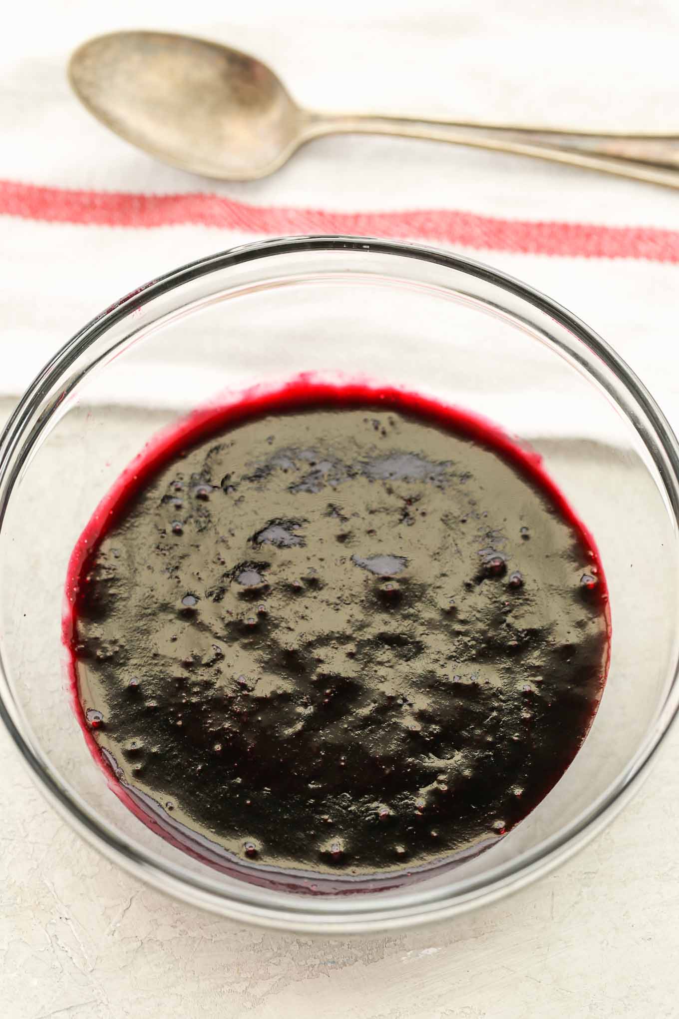 Blueberry sauce for cheesecake in a glass bowl. A spoon and striped towel are behind it. 