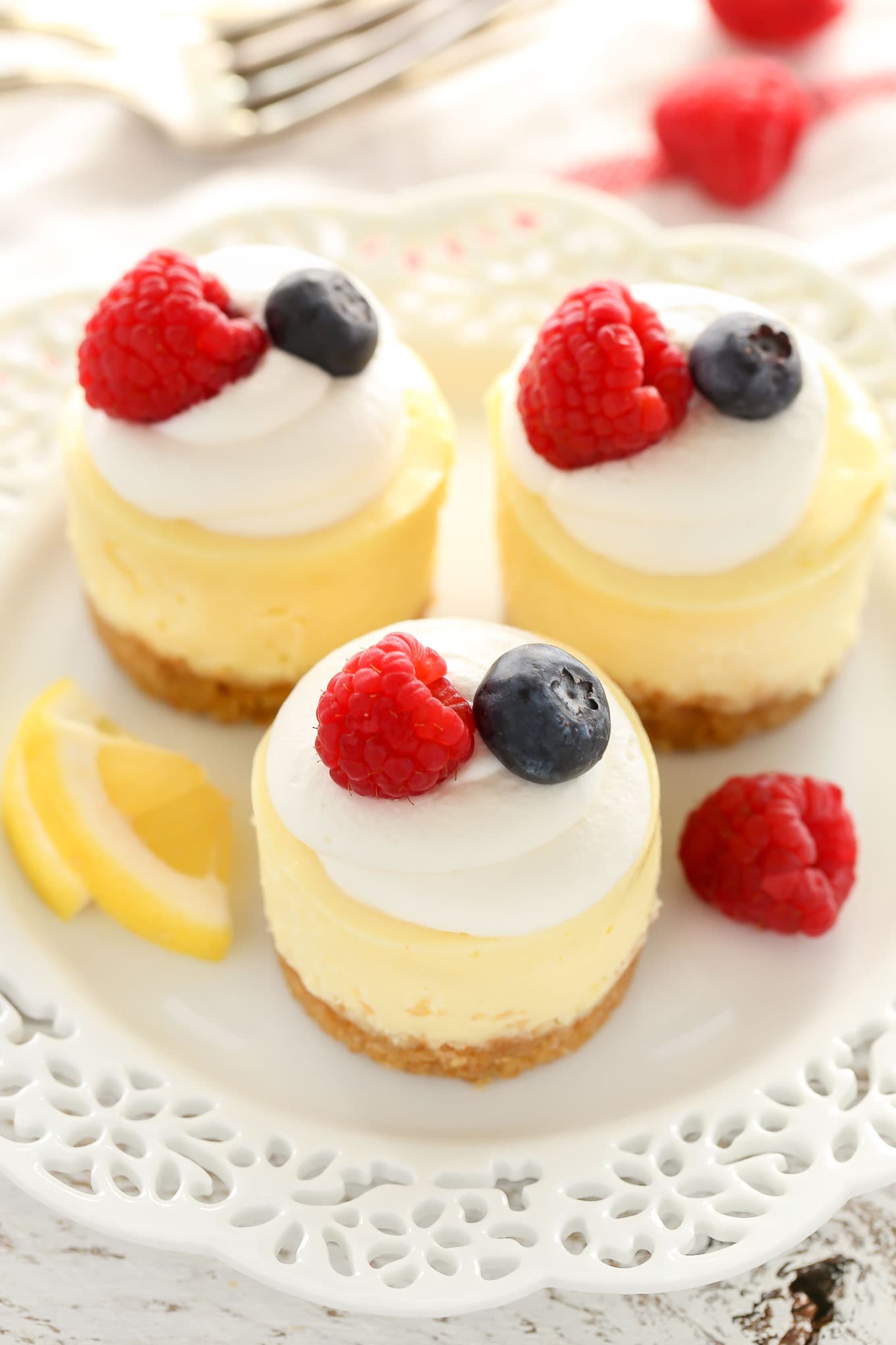 Overhead view of three mini lemon cheesecakes garnished with whipped cream and berries on a white plate. A fork rests in the background. 