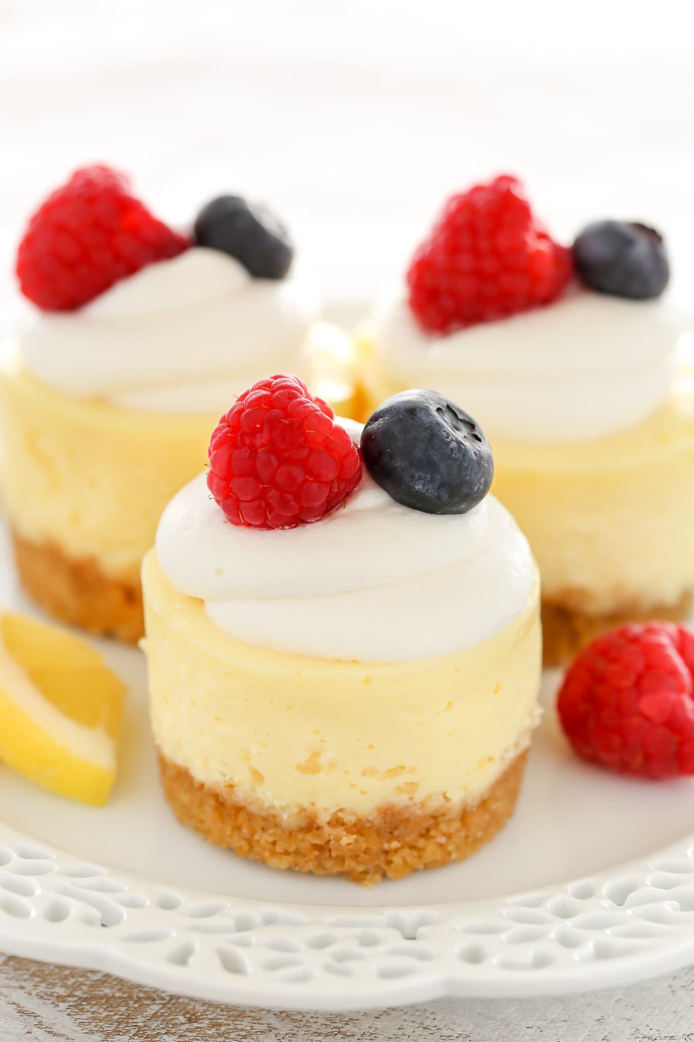 Three mini cheesecakes with a graham cracker crust on a white plate. Each mini cheesecake is garnished with whipped cream and fresh berries. 