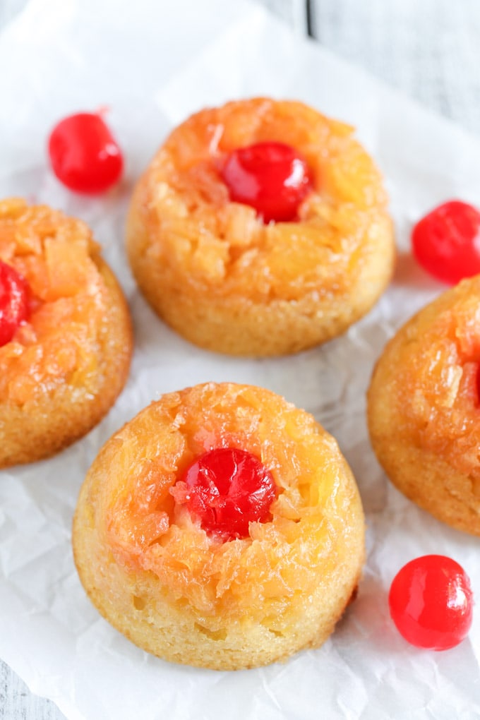 Four pineapple upside down cake cupcakes, surrounded by maraschino cherries, on a piece of parchment paper. 