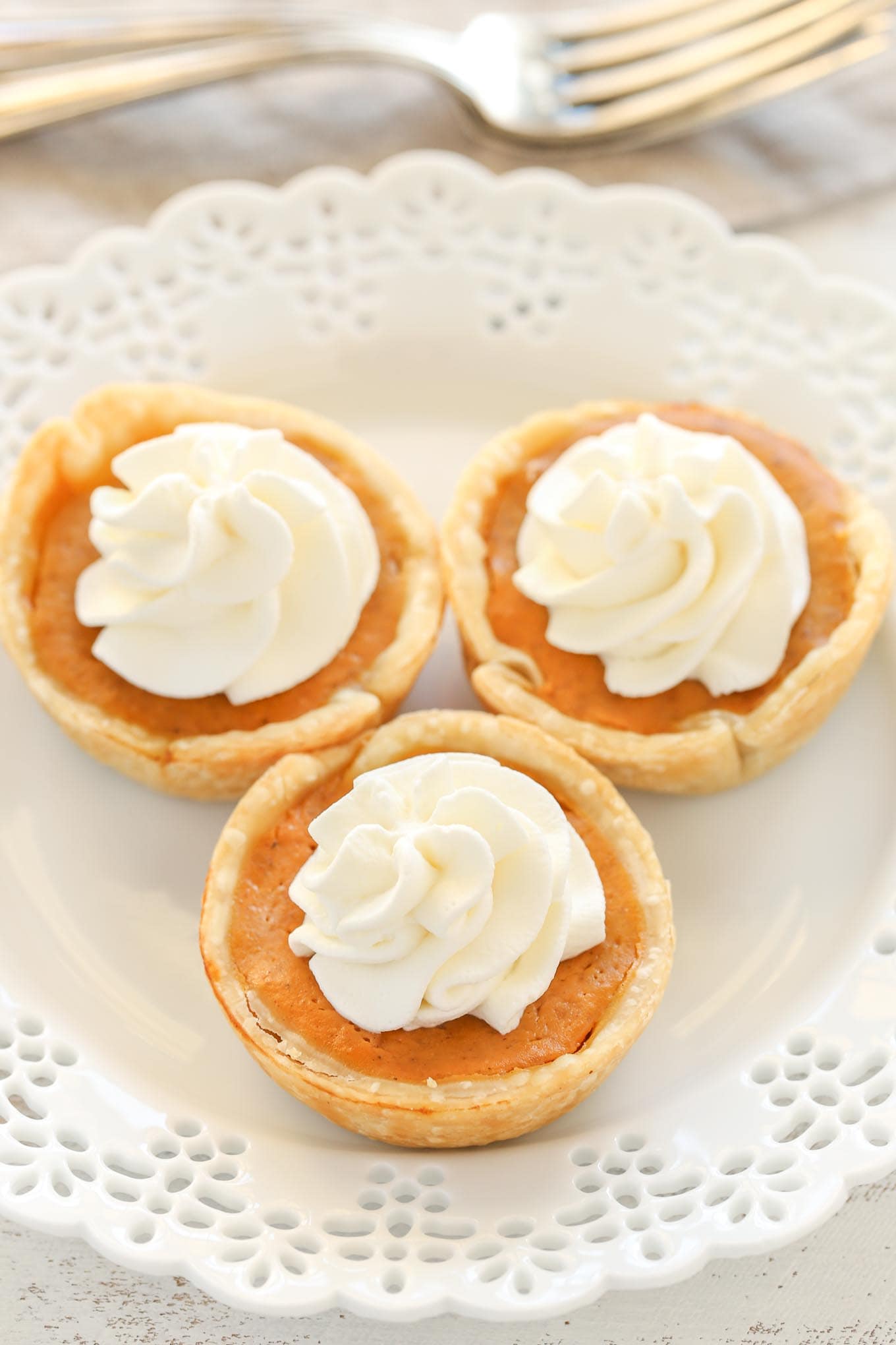 An angled overhead view of three mini pumpkin pies topped with whipped cream on a white plate. Two forks rest in the background.