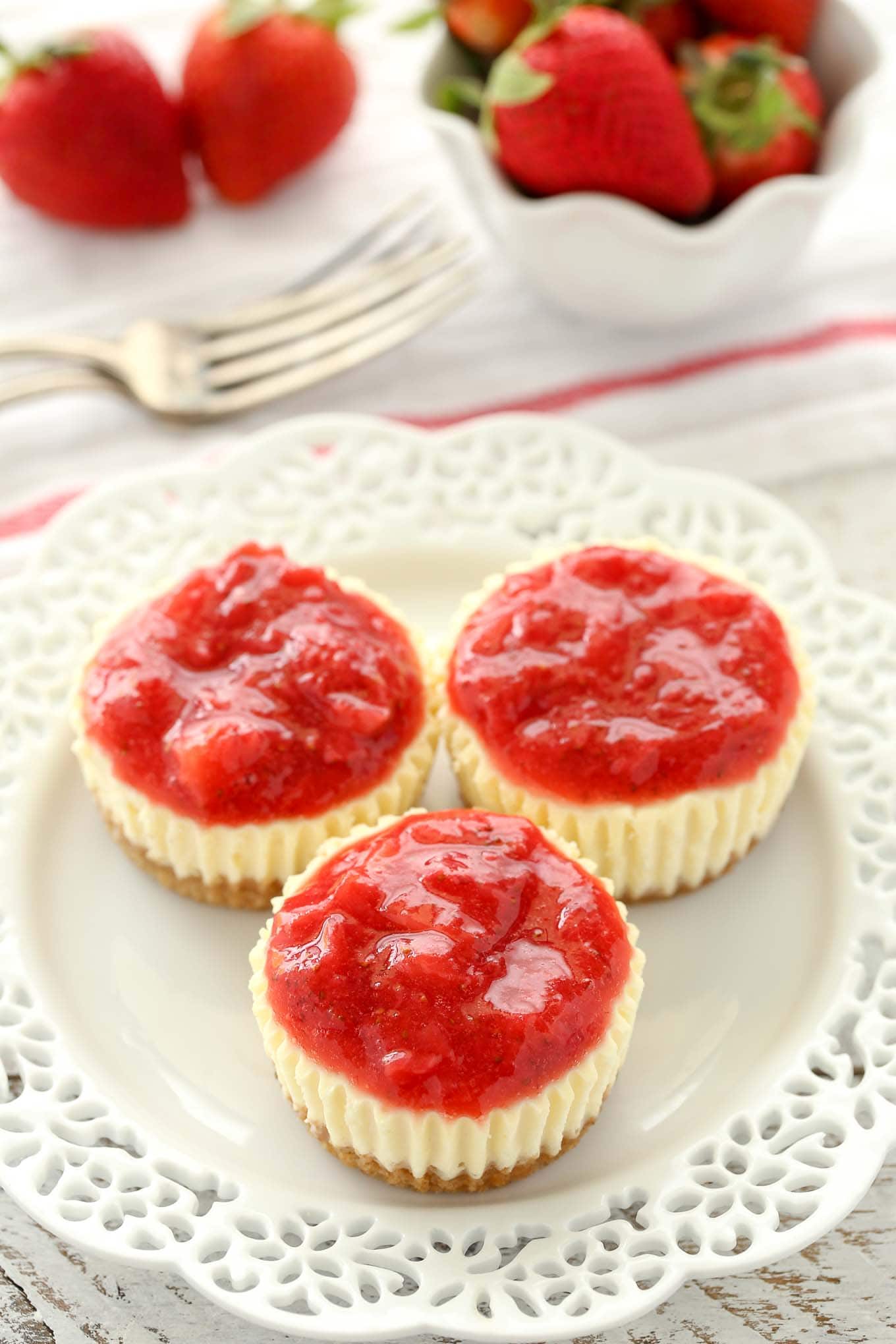 Three homemade strawberry cheesecake bites on a white plate. A fork and a small bowl of fresh strawberries rests in the background. 