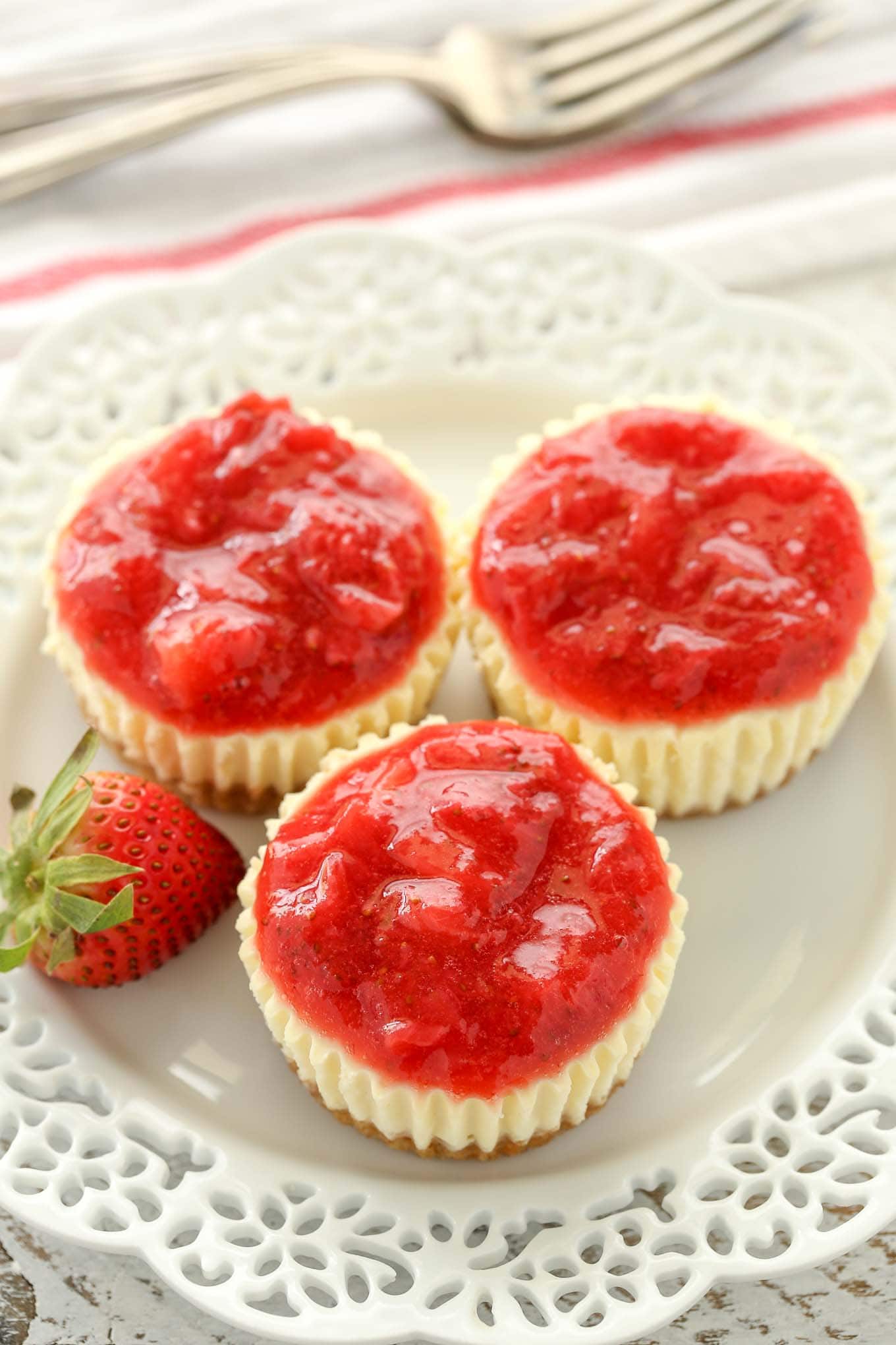 Three mini strawberry cheesecakes on a white plate alongside a fresh strawberry. A fork rests in the background. 