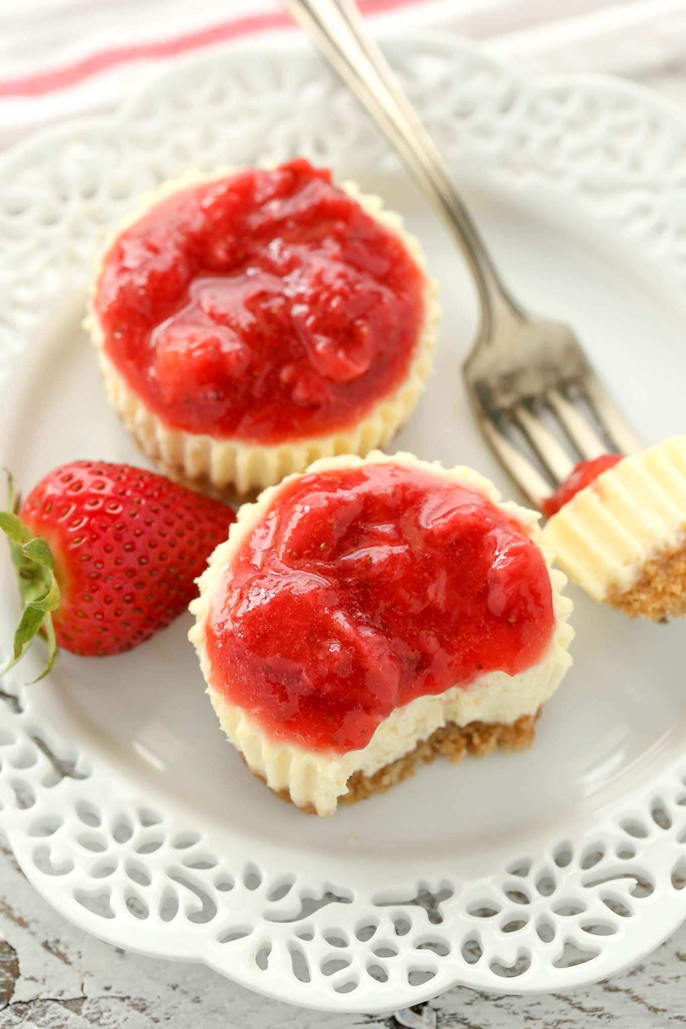 Two strawberry cheesecake bites on a white plate. One mini cheesecake is missing a bite, which is speared on a fork that's also sitting on the plate.