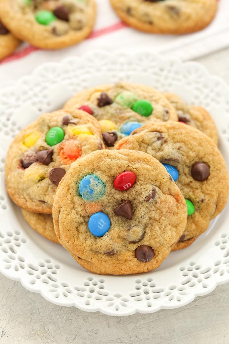 A pile of M&M's cookies on a white plate. Additional cookies rest in the background. 