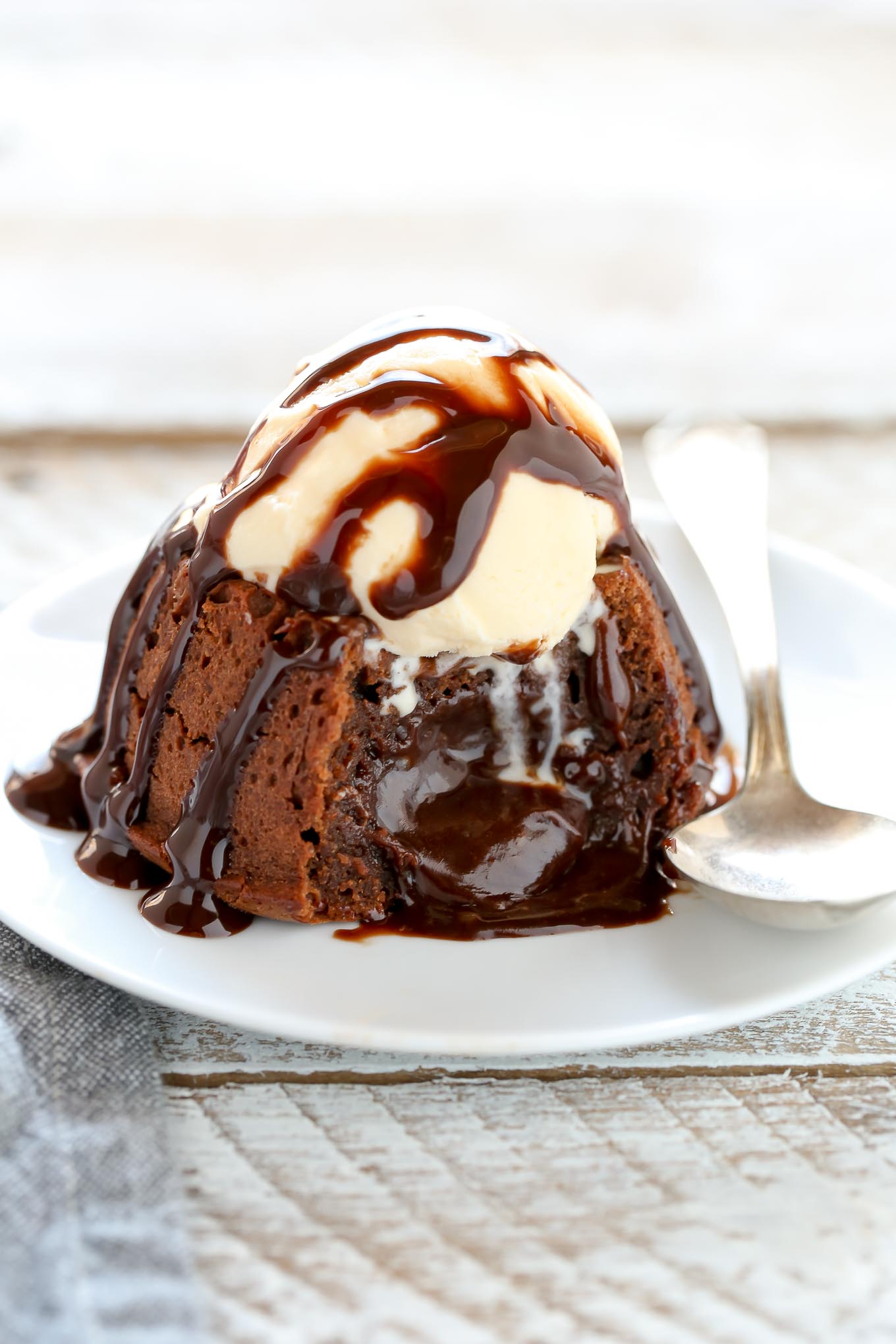 A molten chocolate cake topped with ice cream and chocolate sauce on a white plate. The cake has been cut into and the filling is oozing out. 