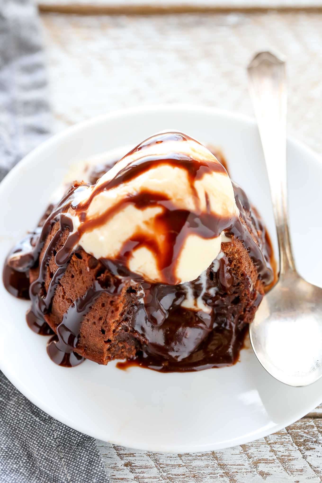 A molten lava cake topped with ice cream and chocolate sauce on a white plate. The cake has been cut into and the filling is oozing out. 