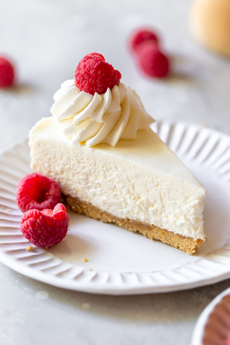 A single slice of no-bake cheesecake on a white plate topped with whipped cream and raspberries.