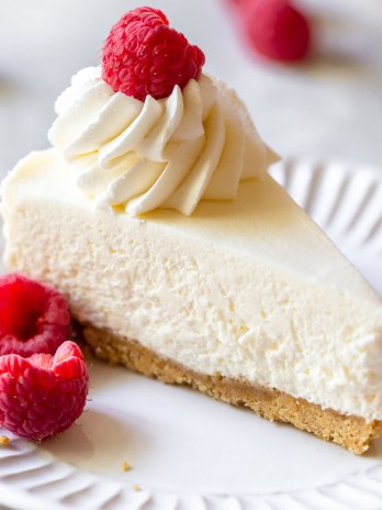 A single slice of no-bake cheesecake on a white plate topped with whipped cream and fresh raspberries.