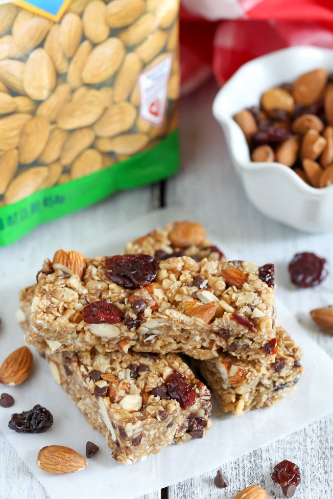 Three no bake granola bars on a square of parchment paper. A bag and a dish of almonds rest in the background. 