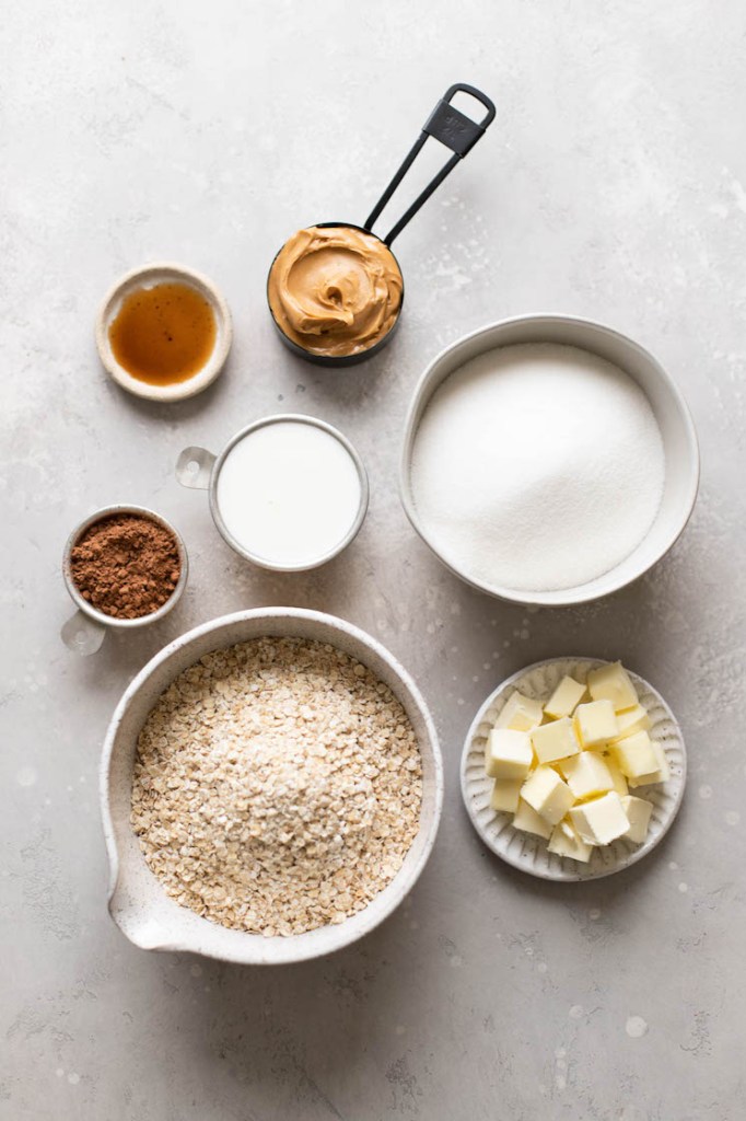 The ingredients needed to make classic no-bake cookies in different bowls and measuring cups on a gray surface.