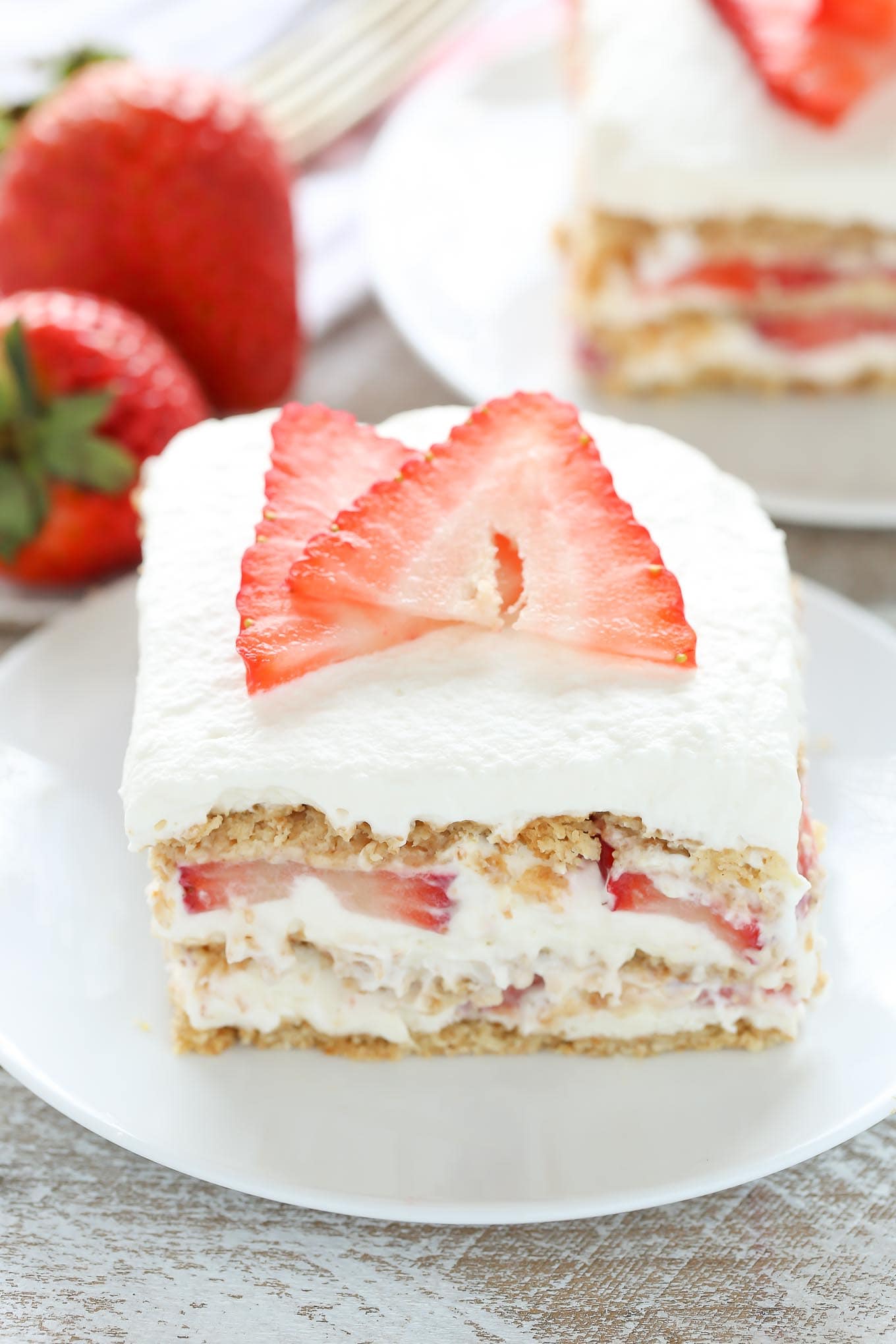 Close up of a slice of strawberry cream cheese icebox cake on a white dessert plate. Another slice and some fresh berries rest in the background. 