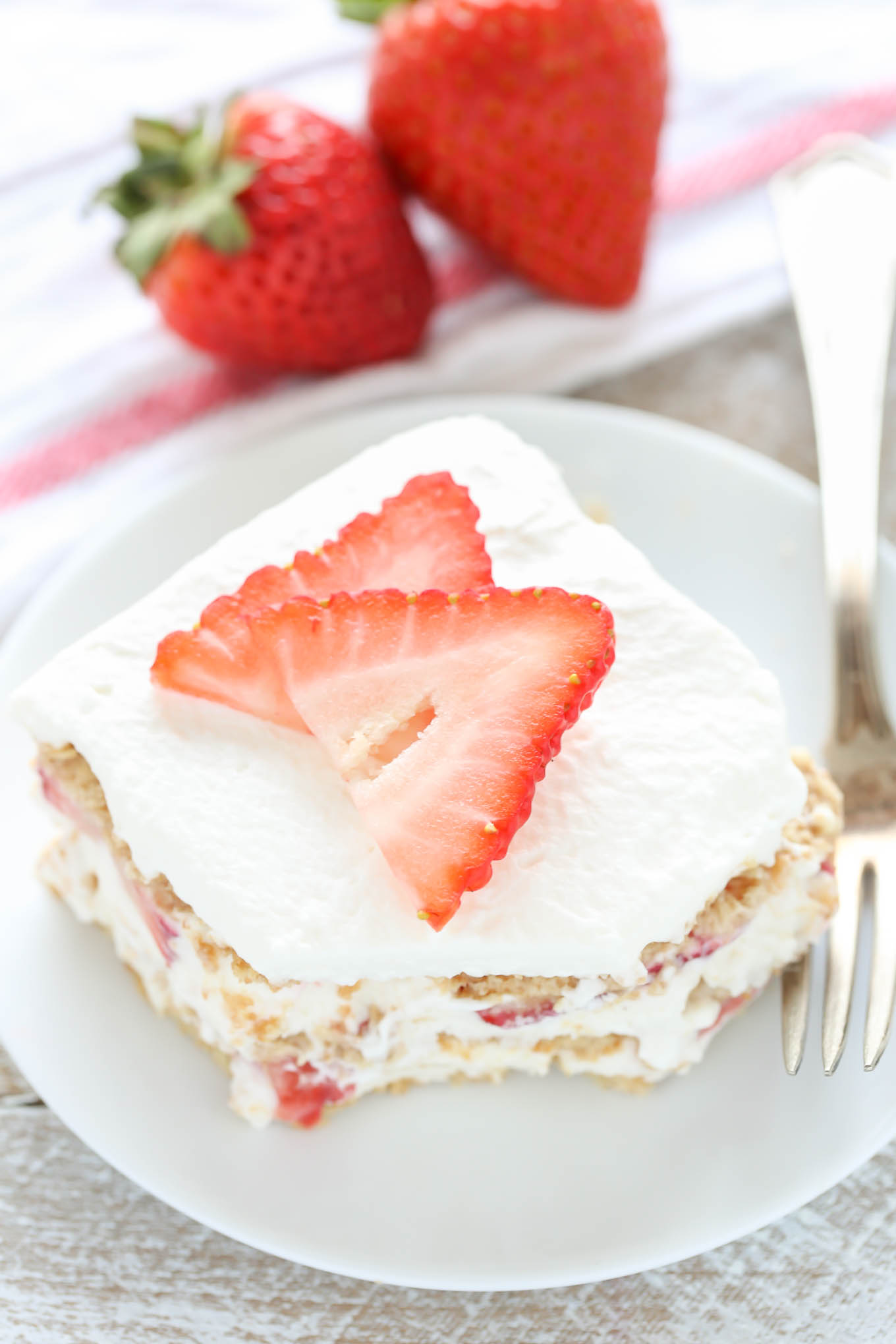 A slice of strawberry graham cracker icebox cake on a white plate with a fork. Two fresh berries rest in the background on a towel. 