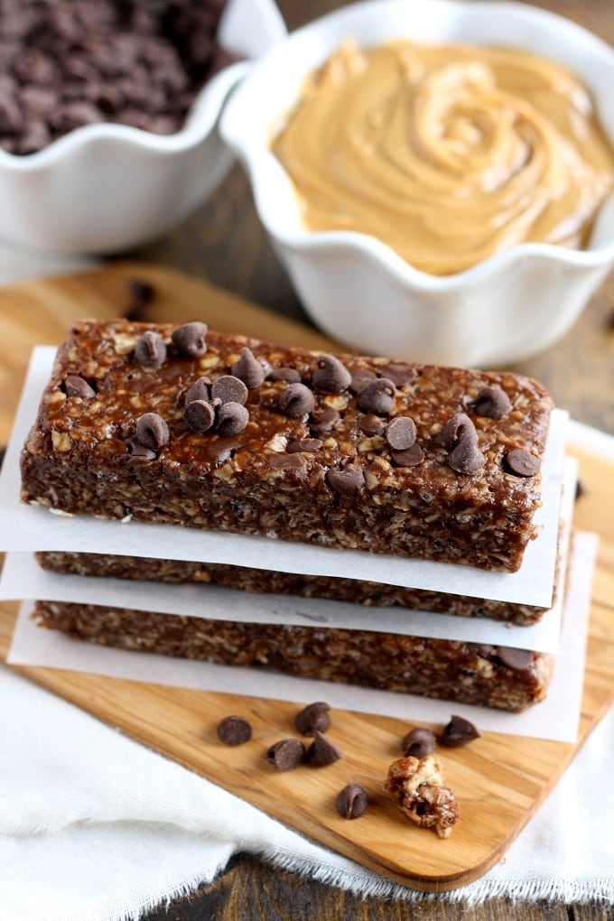 A stack of three peanut butter chocolate chip granola bars on pieces of parchment paper. A bowl of chocolate chips and a bowl of peanut butter rest in the background. 