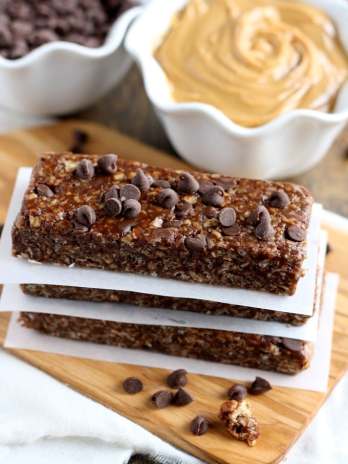 A stack of three peanut butter chocolate chip granola bars on pieces of parchment paper. A bowl of chocolate chips and a bowl of peanut butter rest in the background.