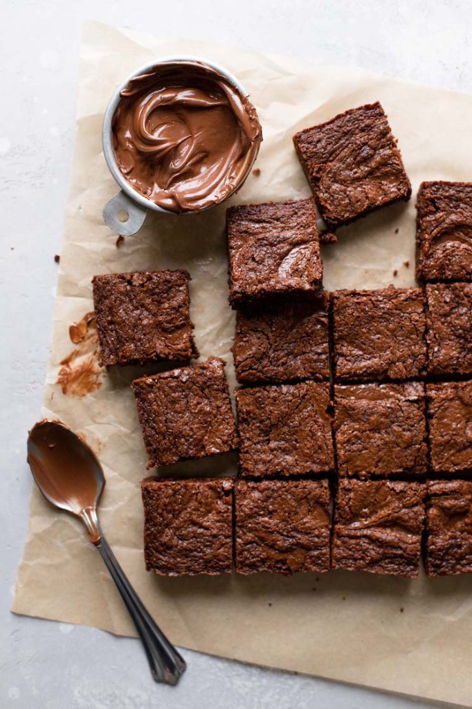 An overhead view of sliced Nutella brownies and a dish of Nutella on a square of parchment paper. A spoon rests near the brownies. 