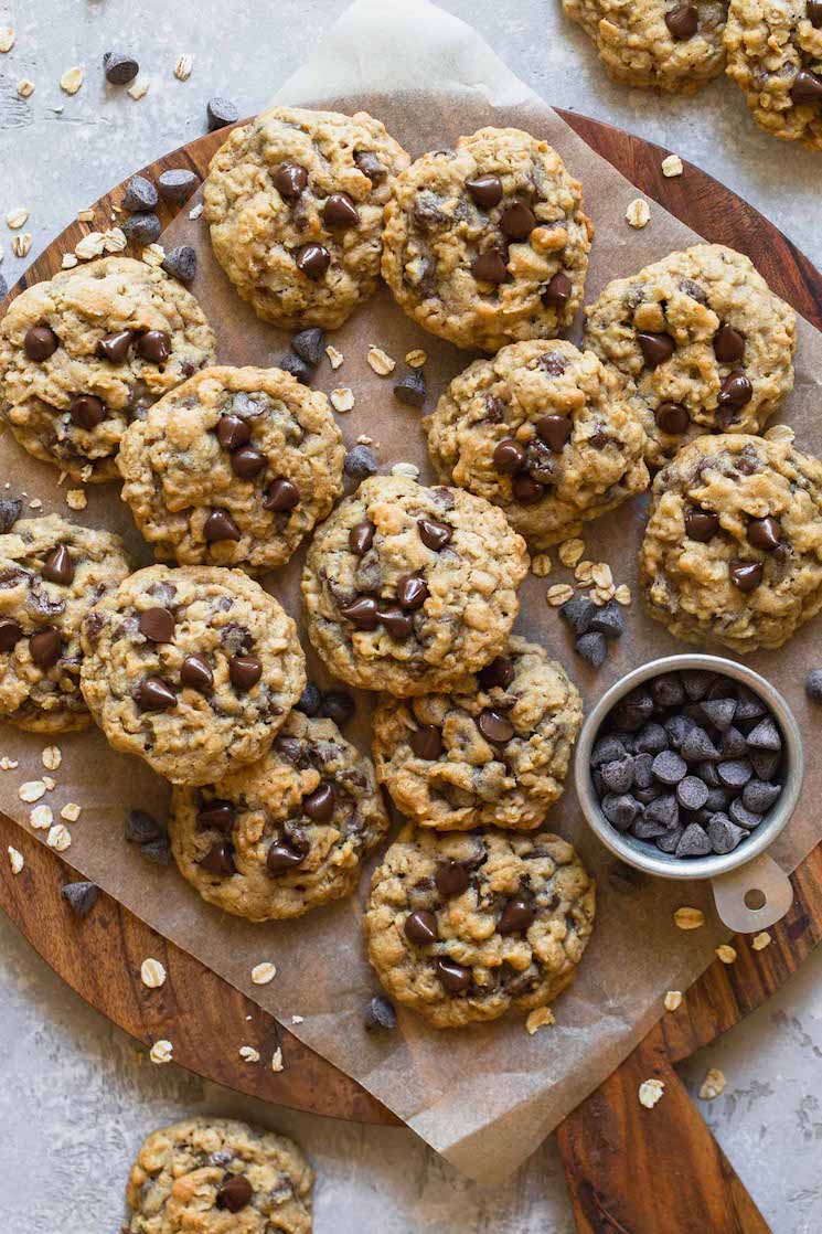 A wooden tray with oatmeal chocolate chip cookies on parchment paper with oats and chocolate chips scattered around them.