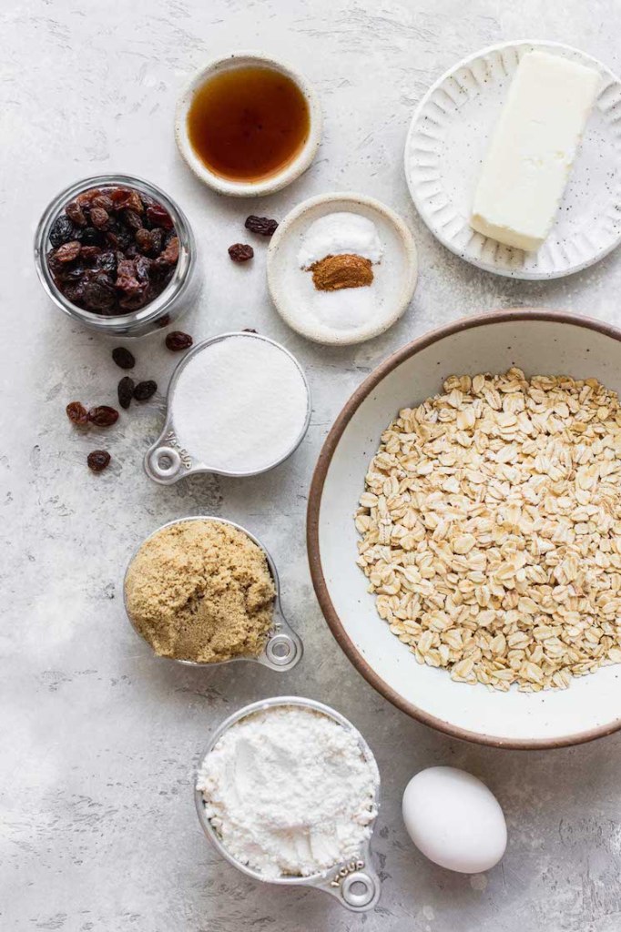 The ingredients needed to make homemade oatmeal raisin cookies laid out in bowls and plates on top of a gray surface.