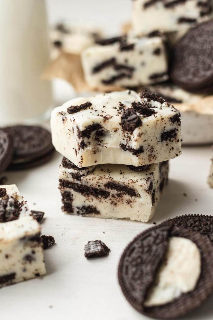 Two pieces of Oreo no bake fudge in a stack. Additional fudge slices and Oreo cookies are scattered around the stack. 