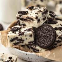 Several pieces of Oreo fudge in a bowl lined with brown parchment paper. More pieces of fudge and a jug of milk surround the bowl.