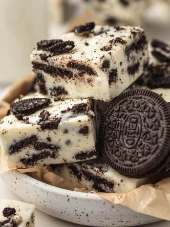 Several pieces of Oreo fudge in a bowl lined with brown parchment paper. More pieces of fudge and a jug of milk surround the bowl.